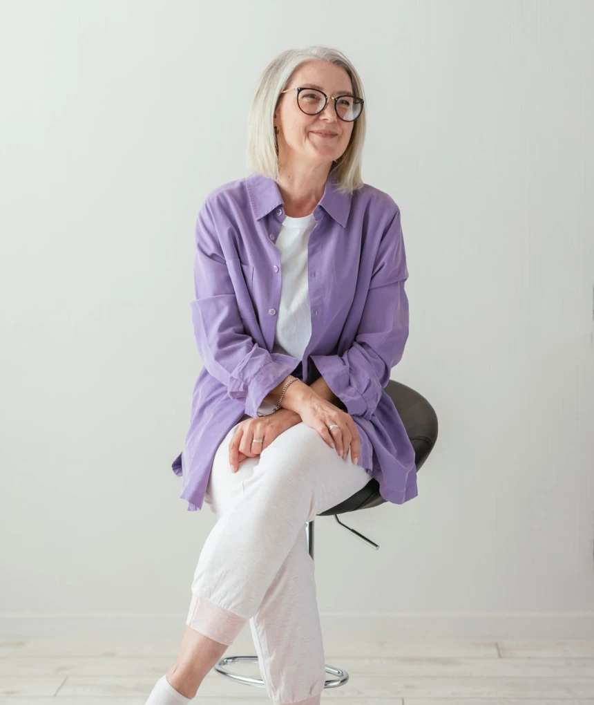 Elderly woman in a purple shirt and white pants, sitting on a stool in a white, neutral room, looking to the right.